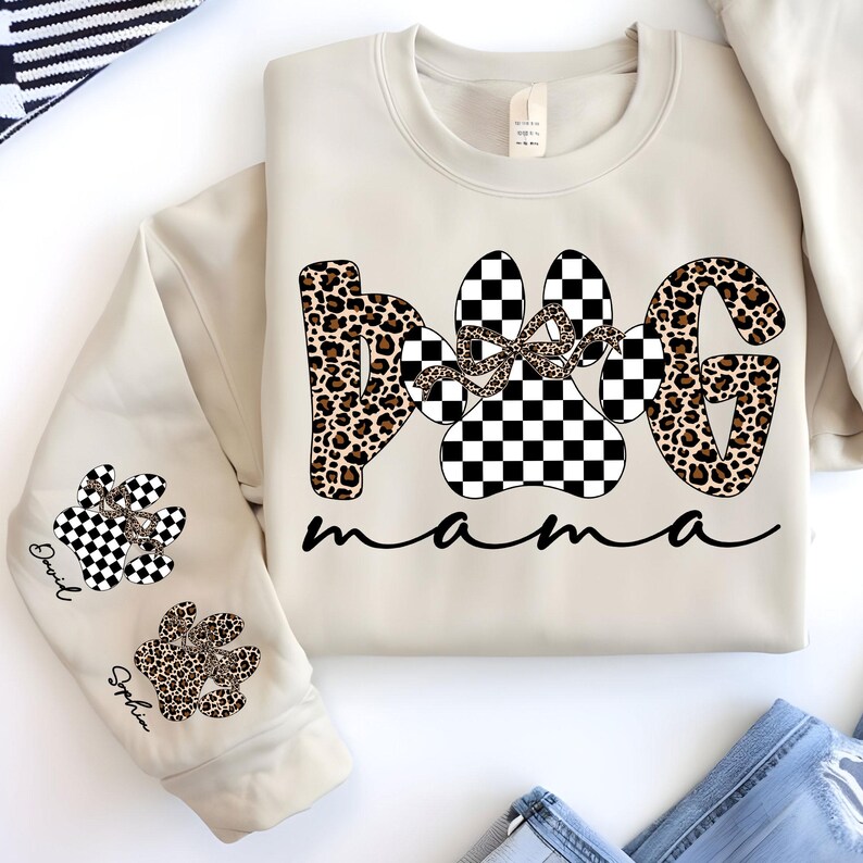 May include: Beige sweatshirt with the words "Dog Mama" in a mix of leopard print and checkered paw print designs. The sleeve features paw prints with names. A bow is in the center of the paw print design.