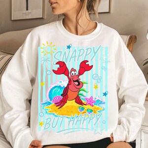 May include: White sweatshirt with a colorful graphic featuring a cartoon crab with the text "SNAPPY BUT HAPPY". The crab is depicted on a beach with a wave, sunglasses, and starfish. The design includes a sun, palm tree, and bubbles.