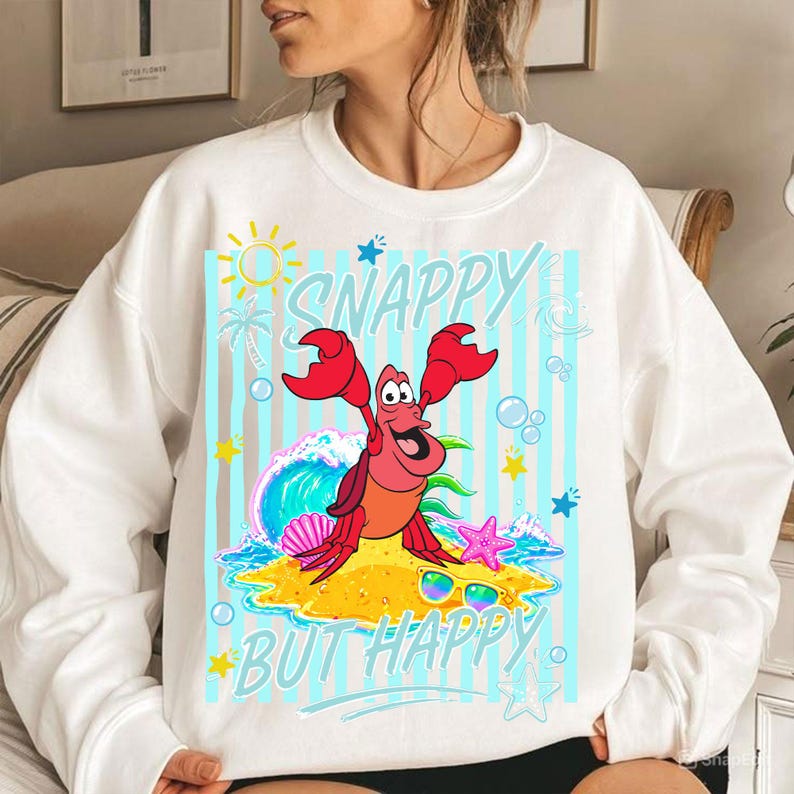 May include: White sweatshirt with a colorful graphic featuring a cartoon crab with the text "SNAPPY BUT HAPPY". The crab is depicted on a beach with a wave, sunglasses, and starfish. The design includes a sun, palm tree, and bubbles.