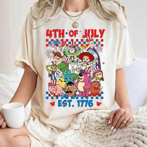 May include: A white t-shirt with a graphic of Toy Story characters celebrating the 4th of July. The text "4th of July" and "Est. 1776" are printed on the shirt.