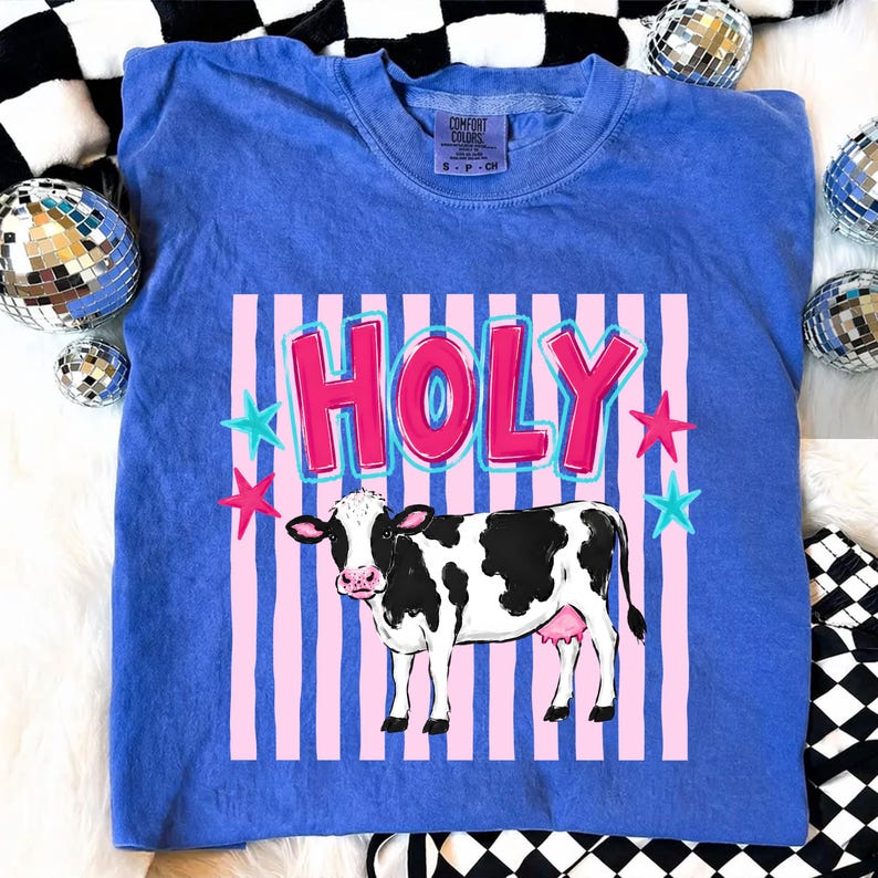 Puede incluir: Camiseta azul Comfort Colors con un gr&aacute;fico de una vaca en blanco y negro y la palabra "HOLY" en letras rosas y azules. El dise&ntilde;o tambi&eacute;n incluye estrellas rosas y azules. La camiseta tiene cuello redondo.