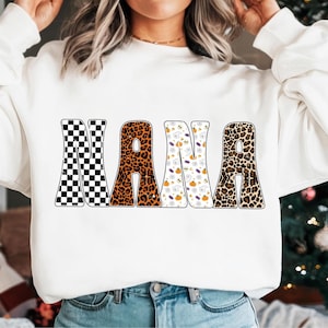 May include: White sweatshirt with the word "NANA" in large, bold letters. The letters have different patterns: black and white checkerboard, leopard print, Halloween-themed pattern with ghosts and pumpkins, and another leopard print.