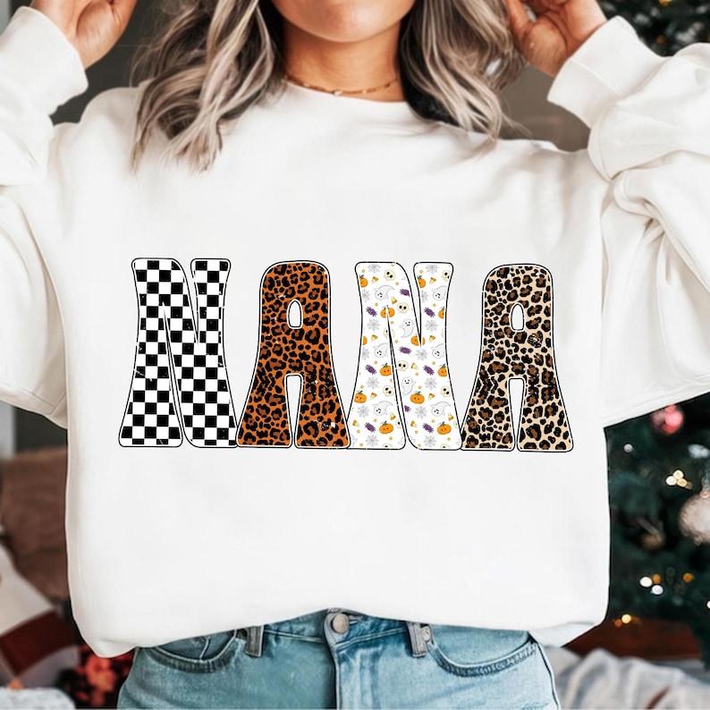 May include: White sweatshirt with the word "NANA" in large, bold letters. The letters have different patterns: black and white checkerboard, leopard print, Halloween-themed pattern with ghosts and pumpkins, and another leopard print.