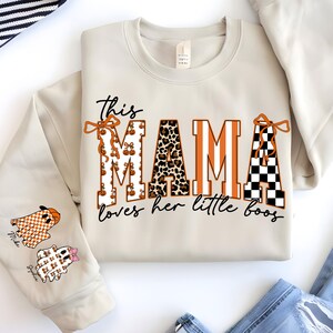 May include: Beige sweatshirt with the text "This MAMA loves her little boos." The word "MAMA" is designed with pumpkin, leopard print, and checkered patterns. Two ghost graphics are on the sleeve.