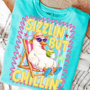 May include: Turquoise crew neck sweatshirt with a graphic of a duck wearing sunglasses and a hat, relaxing in a beach chair while holding a coconut drink. The text reads "Sizzlin' But Chillin'" in yellow and pink.