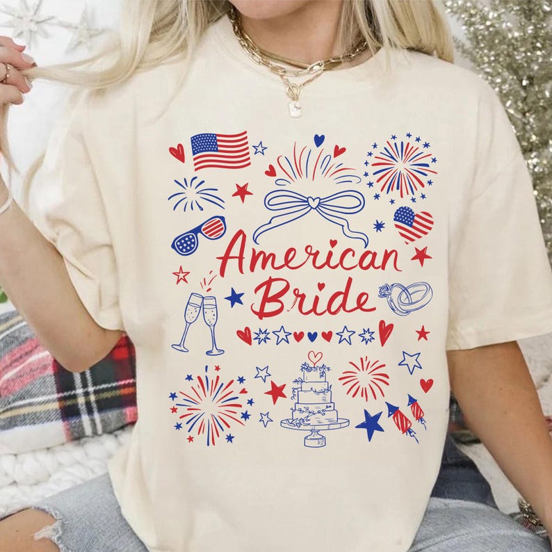 May include: Cream-colored t-shirt with a patriotic design featuring the text "American Bride" in red script. The design includes fireworks, hearts, stars, an American flag, champagne glasses, and a wedding cake.