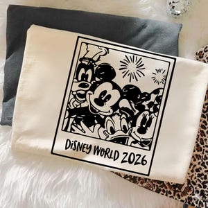 Op de afbeelding: Cr&egrave;mekleurig T-shirt met een zwart-witte afbeelding van Disney-personages, waaronder Mickey Mouse, Minnie Mouse en Donald Duck. De afbeelding is omlijst als een foto met de tekst "DISNEY WORLD 2026". Het shirt is op een witte ondergrond gevouwen.