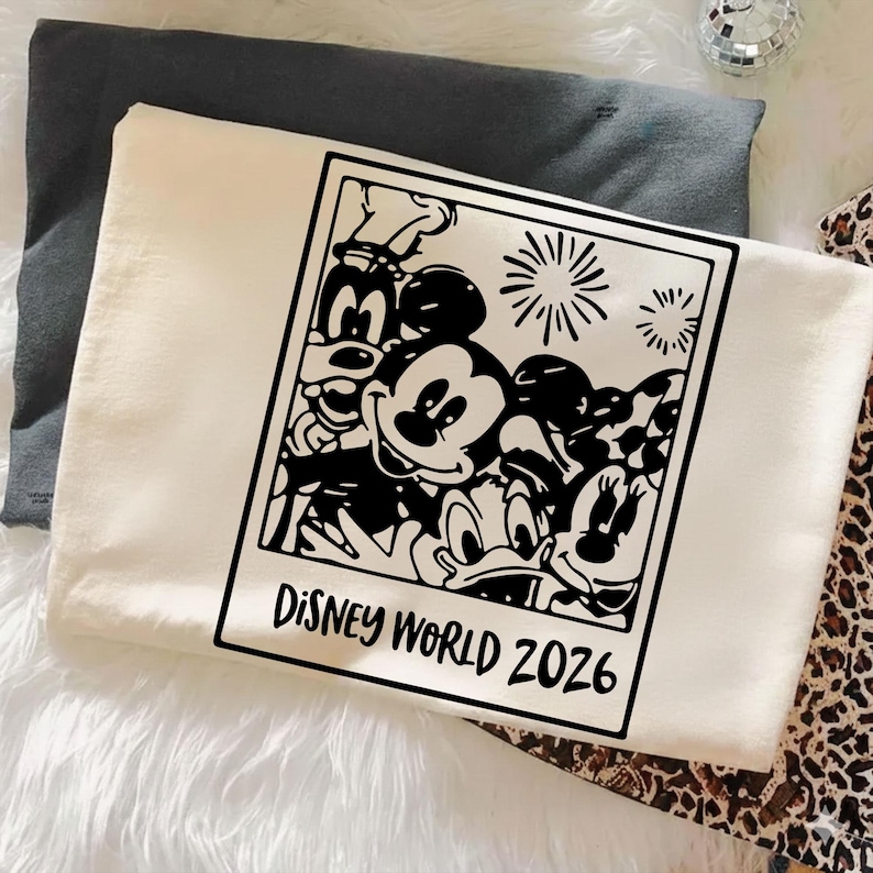 Op de afbeelding: Cr&egrave;mekleurig T-shirt met een zwart-witte afbeelding van Disney-personages, waaronder Mickey Mouse, Minnie Mouse en Donald Duck. De afbeelding is omlijst als een foto met de tekst "DISNEY WORLD 2026". Het shirt is op een witte ondergrond gevouwen.