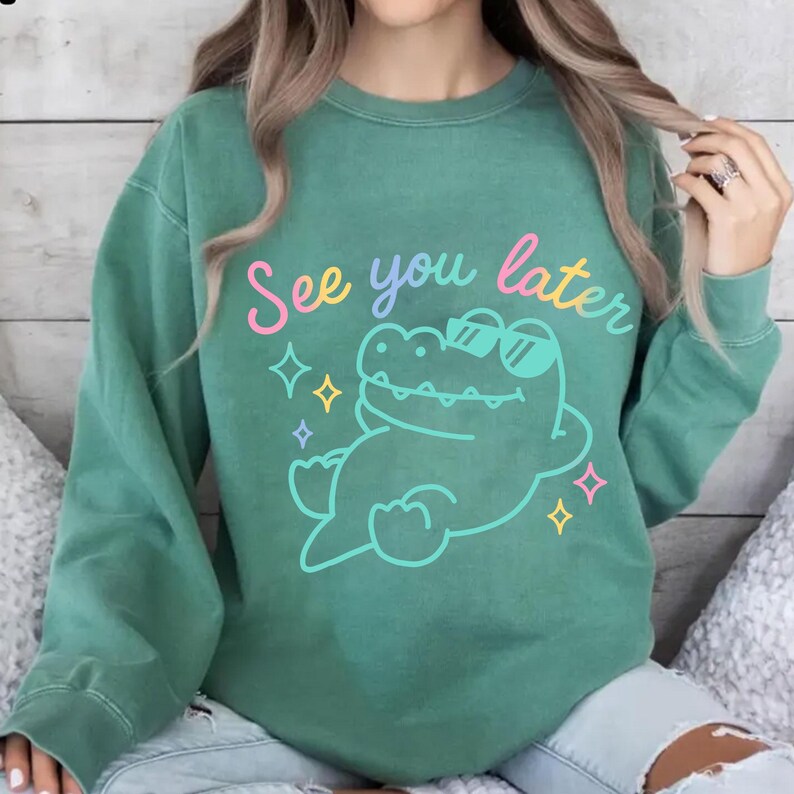 May include: A teal green sweatshirt with the phrase "See you later" in pastel colors. A cartoon alligator wearing sunglasses is below the text, with colorful star accents.