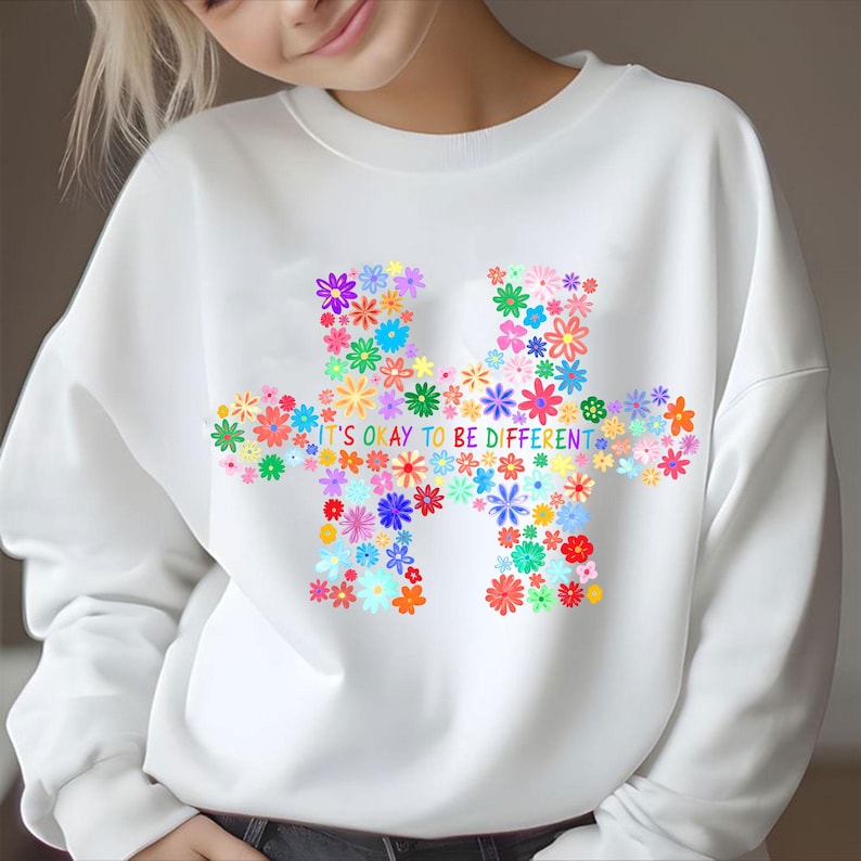 May include: White sweatshirt featuring a colorful floral puzzle piece design. The puzzle is composed of various flowers in shades of red, blue, yellow, and green. The text "IT'S OKAY TO BE DIFFERENT" is printed across the bottom of the puzzle.