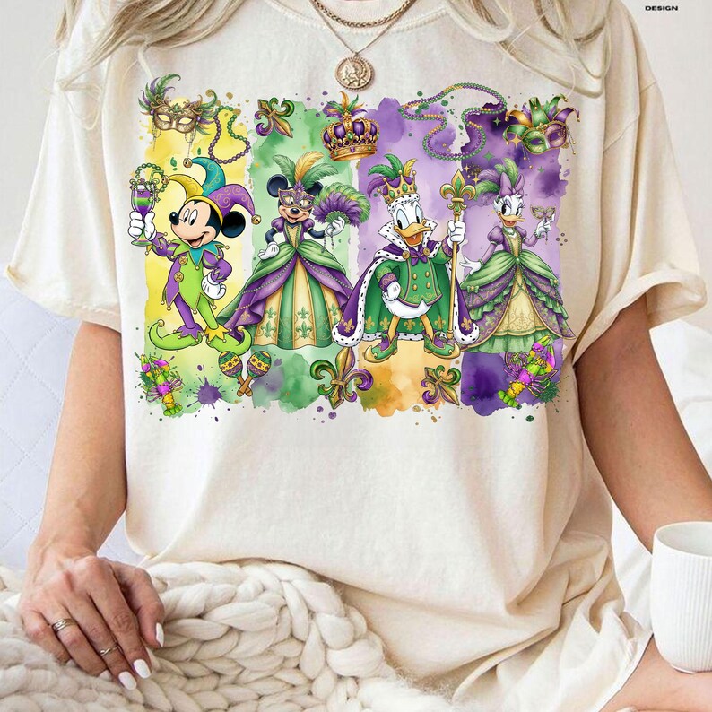 May include: Cream-colored t-shirt featuring a vibrant watercolor design of Disney characters dressed in Mardi Gras attire. Mickey Mouse, Minnie Mouse, Donald Duck, and Daisy Duck are depicted in festive costumes with masks, crowns, and accessories. The design includes fleur-de-lis and beads.