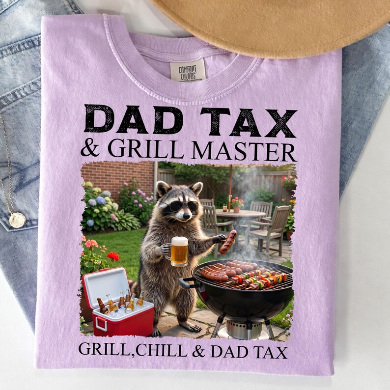 May include: A light purple t-shirt with the text "DAD TAX & GRILL MASTER" in black. A raccoon wearing sunglasses is holding a beer and a sausage near a grill. The text "GRILL, CHILL & DAD TAX" is at the bottom.