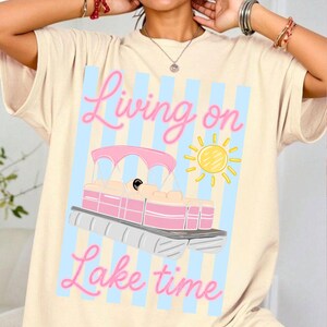 May include: Cream-colored t-shirt featuring a pink and white pontoon boat illustration with a yellow sun. The text "Living on Lake time" is written in pink script above and below the boat design. Blue vertical stripes are in the background.