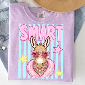 May include: A light purple t-shirt featuring a cartoon donkey wearing heart-shaped sunglasses, a pearl necklace, and a pink fur collar. The word "SMART" is in pink letters above the donkey, with blue vertical stripes in the background.