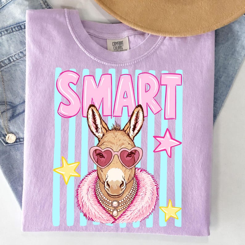 May include: A light purple t-shirt featuring a cartoon donkey wearing heart-shaped sunglasses, a pearl necklace, and a pink fur collar. The word "SMART" is in pink letters above the donkey, with blue vertical stripes in the background.