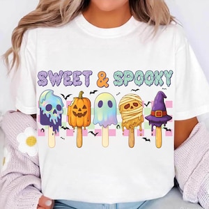 Op de afbeelding: Wit T-shirt met een kleurrijke Halloween-afbeelding. Het ontwerp toont ijslolly-vormige illustraties van een schedel, pompoen, spook, mummie en heksenhoed, met de tekst "SWEET & SPOOKY" erboven. Vleermuizen zijn verspreid.