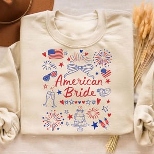 May include: Cream-colored sweatshirt with a festive design featuring the text "American Bride" in red script. The design includes fireworks, American flags, hearts, stars, champagne glasses, and a wedding cake, all in red, white, and blue.