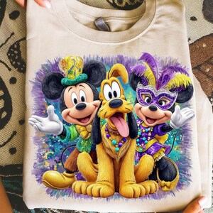 K&ouml;nnte beinhalten: Beiges T-Shirt mit einem farbenfrohen Aufdruck von Mickey Mouse, Pluto und Minnie Mouse in festlicher Kleidung. Mickey tr&auml;gt einen gr&uuml;nen und goldenen Hut, Pluto eine Perlenkette und Minnie eine lila Maske und Federkopfschmuck.
