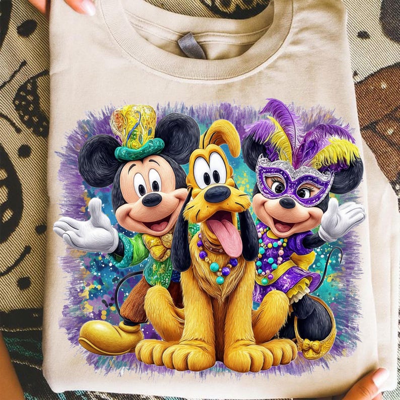 K&ouml;nnte beinhalten: Beiges T-Shirt mit einem farbenfrohen Aufdruck von Mickey Mouse, Pluto und Minnie Mouse in festlicher Kleidung. Mickey tr&auml;gt einen gr&uuml;nen und goldenen Hut, Pluto eine Perlenkette und Minnie eine lila Maske und Federkopfschmuck.