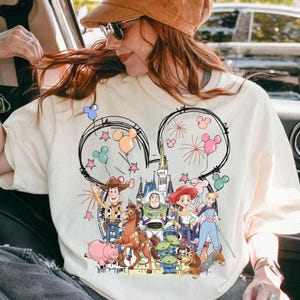 May include: Cream-coloured t-shirt featuring a graphic of Disney's Toy Story characters inside Mickey Mouse ears. The design includes Woody, Buzz Lightyear, Jessie, and other characters, with fireworks and balloons. The shirt is a casual wear item.
