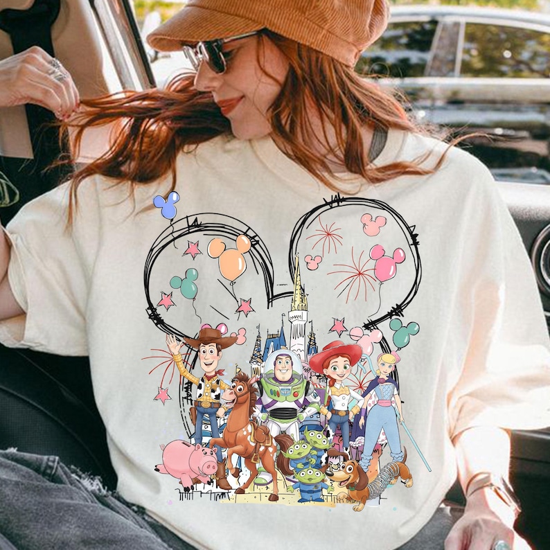 May include: Cream-coloured t-shirt featuring a graphic of Disney's Toy Story characters inside Mickey Mouse ears. The design includes Woody, Buzz Lightyear, Jessie, and other characters, with fireworks and balloons. The shirt is a casual wear item.