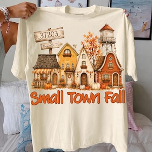May include: A cream-colored t-shirt featuring a graphic of a small town scene with houses, a water tower, pumpkins, and autumn foliage. A wooden sign reads 37203 Nashville, TN. The text Small Town Fall is printed in orange below the houses.