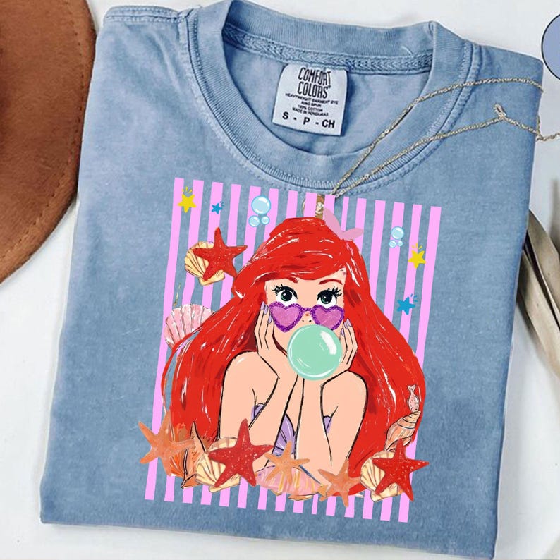 May include: A light blue Comfort Colors t-shirt with a cartoon graphic of a character with red hair, heart-shaped sunglasses, and a bubblegum bubble. The graphic is set against a pink striped background with starfish and seashells.