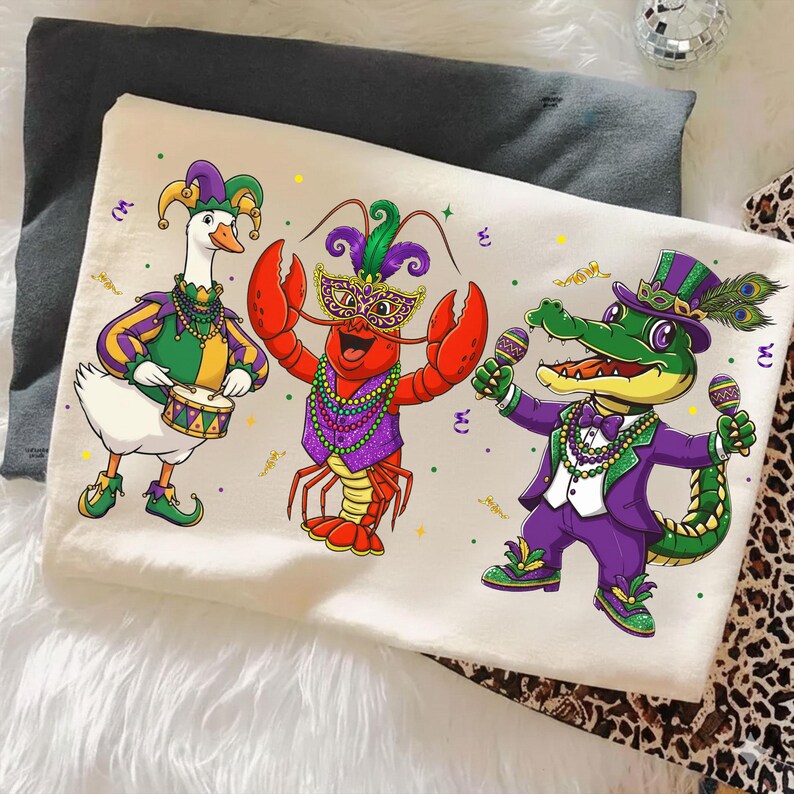 May include: A cream-colored t-shirt featuring cartoon characters dressed in Mardi Gras attire. A goose plays a drum, a lobster wears a mask, and an alligator holds maracas. The characters are in purple, green, and gold.