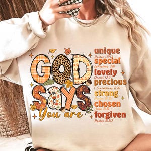 May include: A cream-coloured sweatshirt featuring the words "GOD SAYS You are" in a seasonal design. The word "GOD" has a plaid and leopard print pattern. Additional text includes "unique, special, lovely, precious, strong, chosen, forgiven."