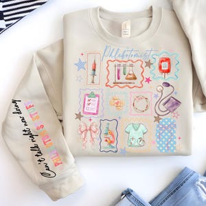 Puede incluir: Sudadera beige con gráficos coloridos de temática médica, que incluyen una jeringa, una bolsa de sangre y un estetoscopio. La manga tiene el texto "Can't talk right now doing phlebotomist stuff."