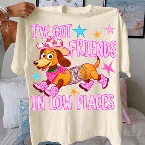 May include: A cream-colored t-shirt featuring a cartoon dachshund with a spring body, wearing a pink cowgirl hat, a pink bandana, and pink cowboy boots. The text reads I'VE GOT FRIENDS IN LOW PLACES, surrounded by colorful star graphics.