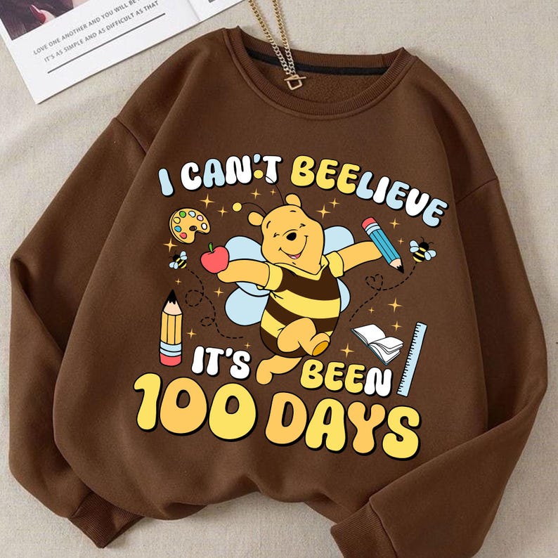 May include: Brown sweatshirt with a graphic of a bear dressed as a bee, with the text "I CAN'T BEELIEVE IT'S BEEN 100 DAYS". The design includes pencils, an apple, a ruler, and a palette. A gold chain is visible at the top.