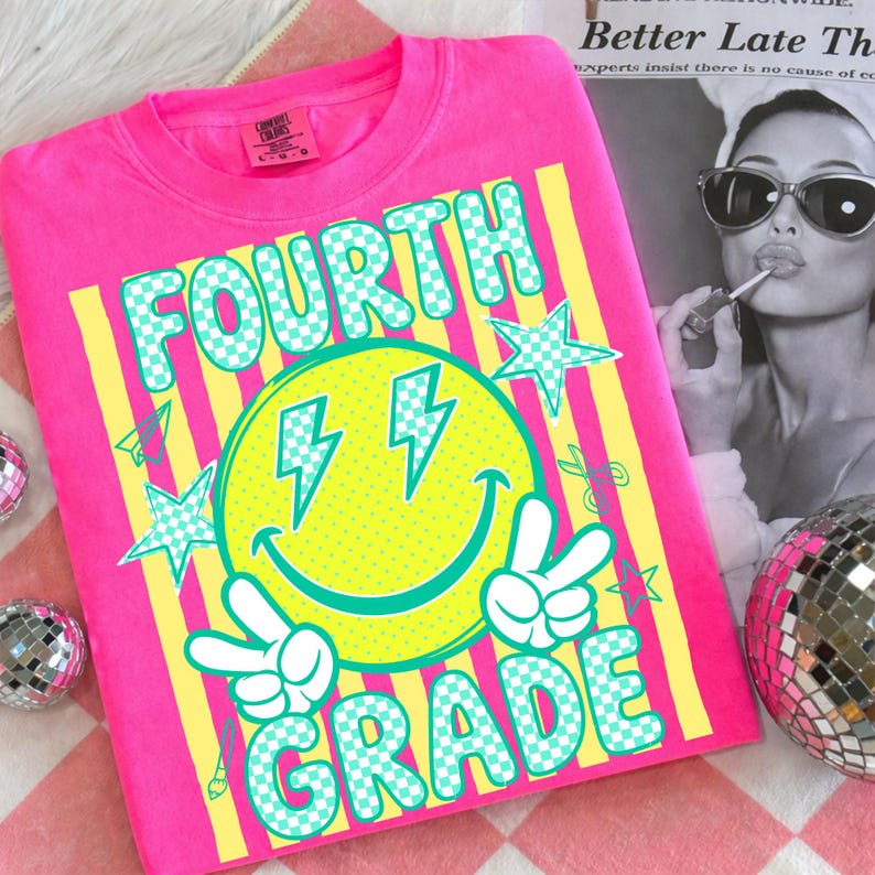 May include: A bright pink Fourth Grade sweatshirt with a large graphic. The graphic features a yellow smiley face with lightning bolt eyes, peace signs, and the words "FOURTH GRADE" in a checkered font. Yellow stripes and stars complete the design.