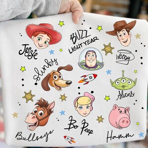 May include: White fabric with colourful illustrations of characters from the animated film "Toy Story." Includes images of Jessie, Buzz Lightyear, Woody, Slinky, Aliens, Bo Peep, Bullseye, and Hamm, with their names written in cursive.