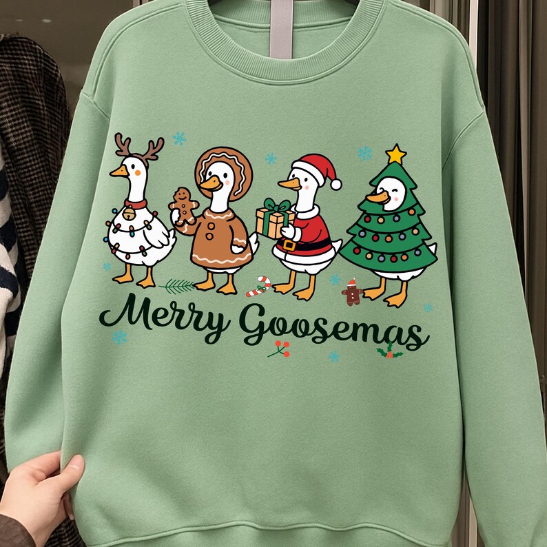 Peut inclure: Un sweat-shirt vert clair avec un motif festif "Merry Goosemas". Le graphisme pr&eacute;sente quatre oies de dessin anim&eacute; v&ecirc;tues de tenues de vacances, dont un renne, un pain d'&eacute;pices, un P&egrave;re No&euml;l et un sapin de No&euml;l. Le texte "Merry Goosemas" est &eacute;crit en police script.