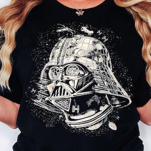Könnte beinhalten: Schwarzes T-Shirt mit einer detaillierten weiß-grauen Grafik von Darth Vaders Helm. Das Design beinhaltet Weltraumelemente und einen Sternenhintergrund, wodurch ein Science-Fiction-Thema entsteht.