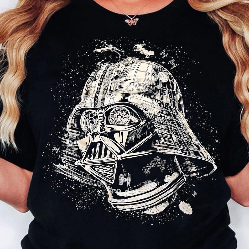 May include: Black t-shirt featuring a detailed graphic of Darth Vader's helmet, rendered in white and gray. The design includes space battle elements and a starry background, creating a sci-fi theme.