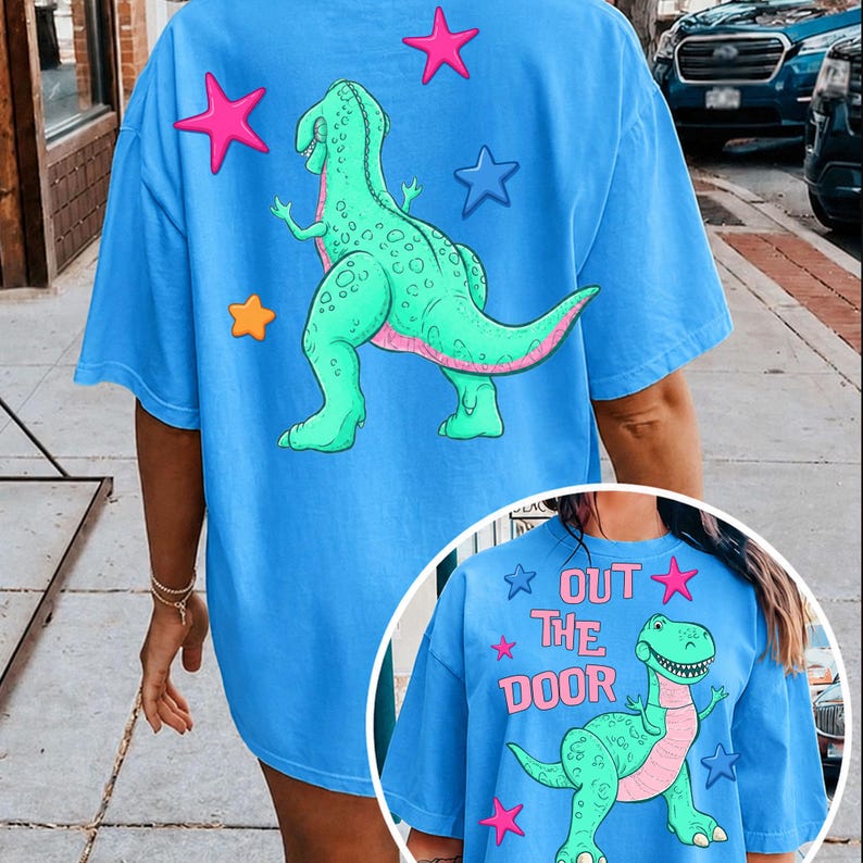Może przedstawiać: Jasnoniebieska koszulka z nadrukiem kresk&oacute;wkowego dinozaura i gwiazd. Dinozaur jest zielony z r&oacute;żowymi detalami. Na koszulce widnieje napis "OUT THE DOOR". Koszulka ma swobodny, obszerny kr&oacute;j.