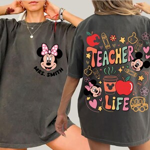 May include: Dark grey t-shirt with Minnie Mouse and a pink bow on the front, along with the name "Mrs. Smith". The back displays "Teacher Life" with colourful graphics like apples, crayons, and Mickey Mouse.