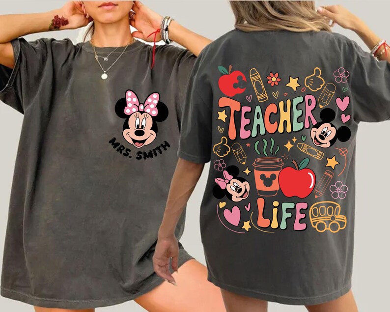 May include: Dark grey t-shirt with Minnie Mouse and a pink bow on the front, along with the name "Mrs. Smith". The back displays "Teacher Life" with colourful graphics like apples, crayons, and Mickey Mouse.