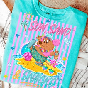 May include: Turquoise crew neck t-shirt with a graphic featuring a cartoon mouse on a beach. The design includes the words "Sun, Sand & Snacks" in pink and blue, along with beach-themed illustrations like a palm tree, sun, and seashells.