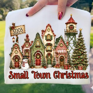 May include: A white t-shirt featuring a festive illustration of a small town during Christmas. The design includes colourful houses, a snowman, a water tower, a Christmas tree, and a sign that reads 37203 Nashville, TN. The text Small Town Christmas is printed in red at the bottom.