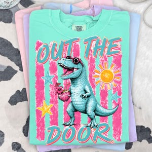 May include: A turquoise t-shirt featuring a blue dinosaur wearing sunglasses and holding a tropical drink. The text "OUT THE DOOR" is above and below the dinosaur. Pink and yellow stars and a sun are also featured.