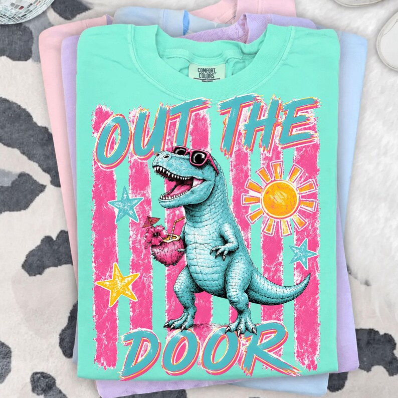 May include: A turquoise t-shirt featuring a blue dinosaur wearing sunglasses and holding a tropical drink. The text "OUT THE DOOR" is above and below the dinosaur. Pink and yellow stars and a sun are also featured.