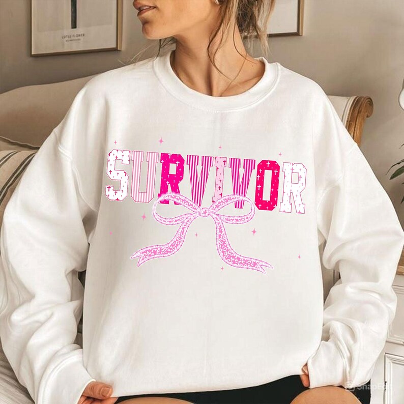 May include: White sweatshirt with the word "SURVIVOR" in pink block letters and a pink ribbon graphic. The letters have various patterns, including stripes and crosses. The sweatshirt is a casual style.