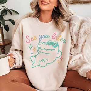May include: Cream-colored sweatshirt with the phrase "See you later" in pastel colors. The graphic features a cartoon alligator wearing sunglasses. The alligator is outlined in turquoise, with a white body and sunglasses. The design includes colorful star accents.