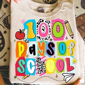 May include: A beige t-shirt with a colorful graphic that reads "100 Days of School." The design includes an apple, pencil, and other school-themed elements. The text is in various bright colors and fonts.