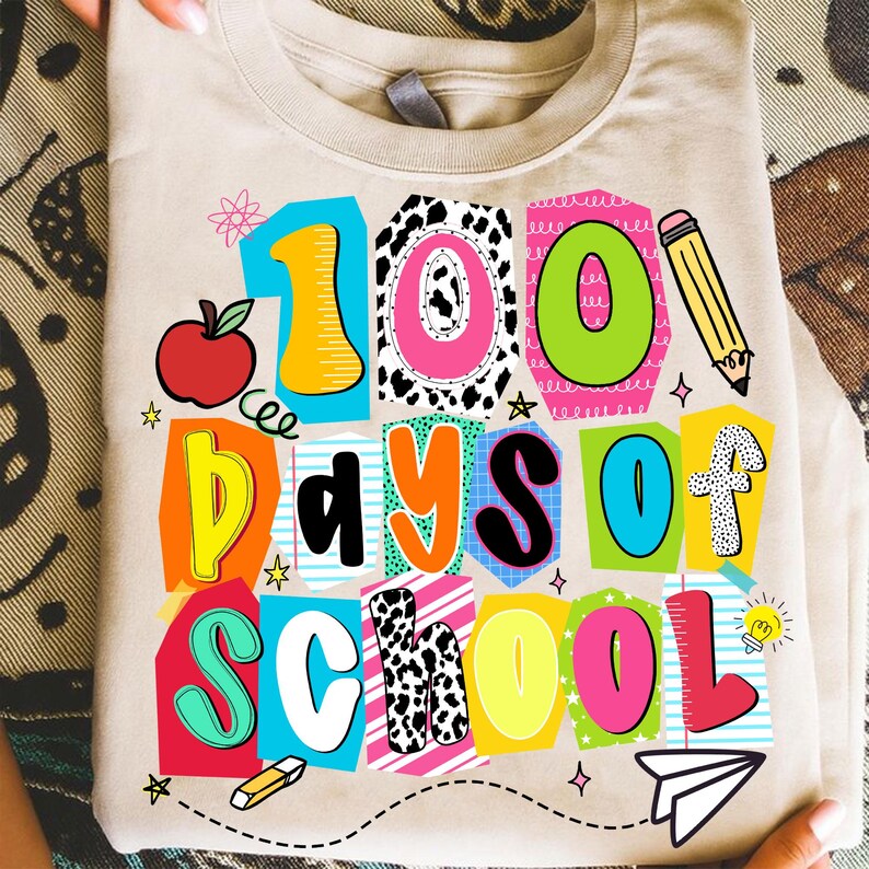 May include: A beige t-shirt with a colorful graphic that reads "100 Days of School." The design includes an apple, pencil, and other school-themed elements. The text is in various bright colors and fonts.