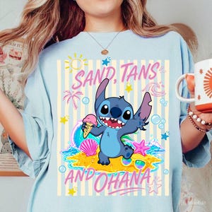 Może przedstawiać: Jasnoniebieska koszulka z postacią z kresk&oacute;wki trzymającą lody, ze słowami "SAND, TANS AND OHANA". Projekt zawiera scenę plażową z palmami, falami i gwiazdami.