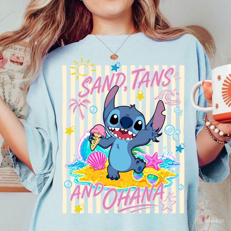 Może przedstawiać: Jasnoniebieska koszulka z postacią z kresk&oacute;wki trzymającą lody, ze słowami "SAND, TANS AND OHANA". Projekt zawiera scenę plażową z palmami, falami i gwiazdami.